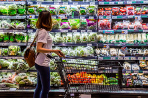 Woman with empty shopping cart in produce section of grocery store. Most items are packaged in plastic.